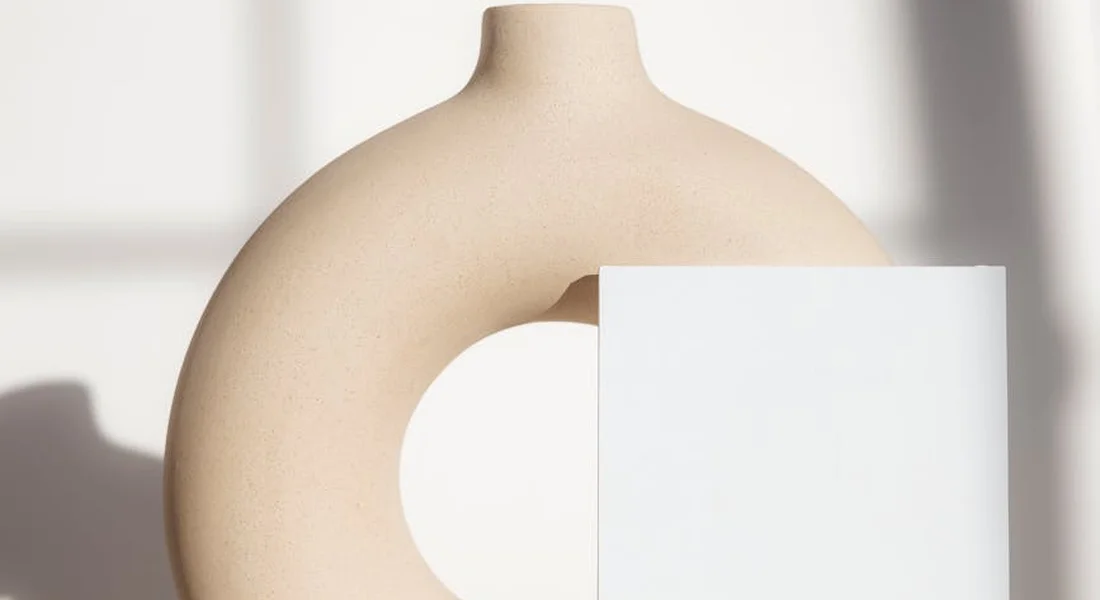 Abstract ceramic sculpture with a curved, pale beige surface and a circular cutout, partially overlapped by a white rectangular panel against a light background.