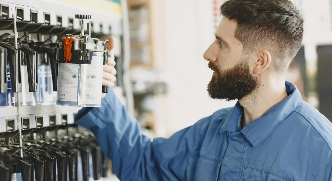 Bearded technician in a blue shirt selecting aerosol spray cans from a wall of automotive paints in a shop.