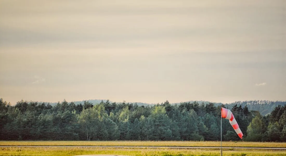 Wide view of an airfield with a windsock, open field, and a tree line on the horizon under a pale sky.