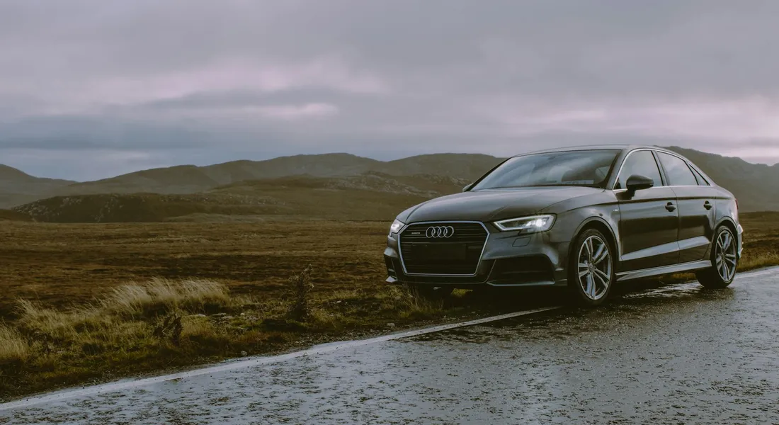 Dark gray Audi sedan on a wet road in a moorland setting with rolling hills and an overcast sky.