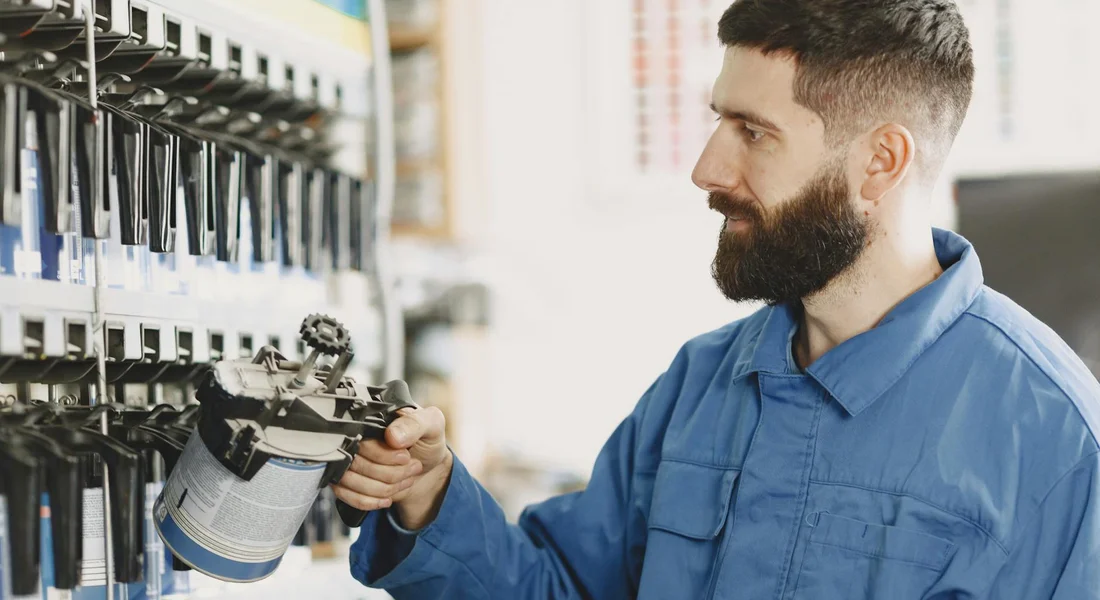 Bearded technician in a blue uniform holding a chemical spray container while inspecting a wall of spray nozzles in a workshop.