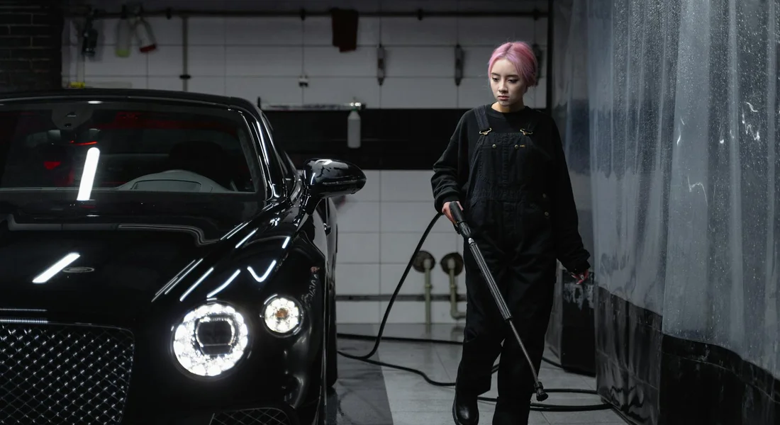A person with pink hair wearing black clothing stands beside a glossy black car in a workshop, holding a detailing tool to lock in the shine.