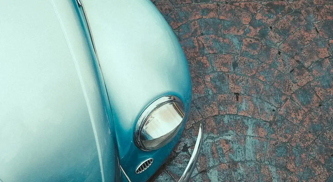 Close-up of a blue vintage car's front end showing a headlight and chrome bumper on a textured pavement.