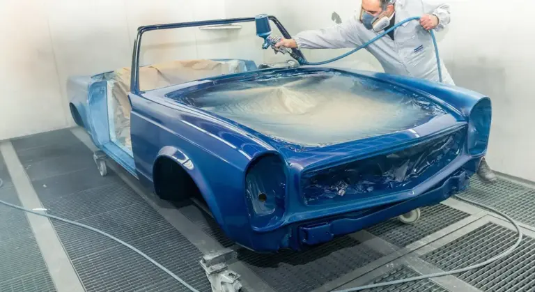 A technician wearing protective gear sprays blue paint onto a classic car body inside a paint booth, with masking film covering windows and interior areas.