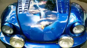 Close-up of a glossy blue Porsche car hood with vented scoops and round headlights reflecting the sky.