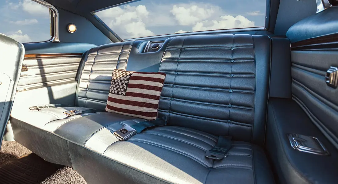 Backseat of a vintage car with blue upholstered seats and a small American flag pillow.