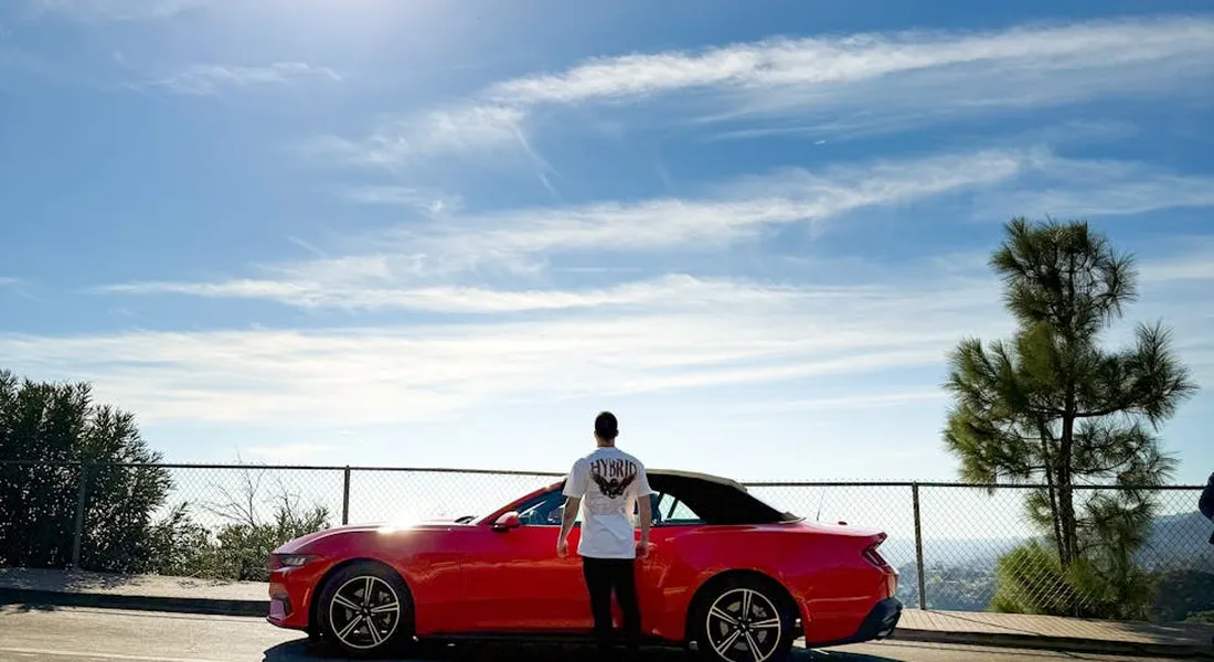 A person stands beside a red convertible on a sunny day in an outdoor parking area with a chain-link fence and clear blue sky.