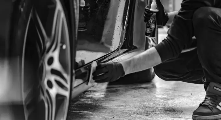 Detailer cleaning the side of a car, using a spray bottle and microfiber cloth near the wheel.
