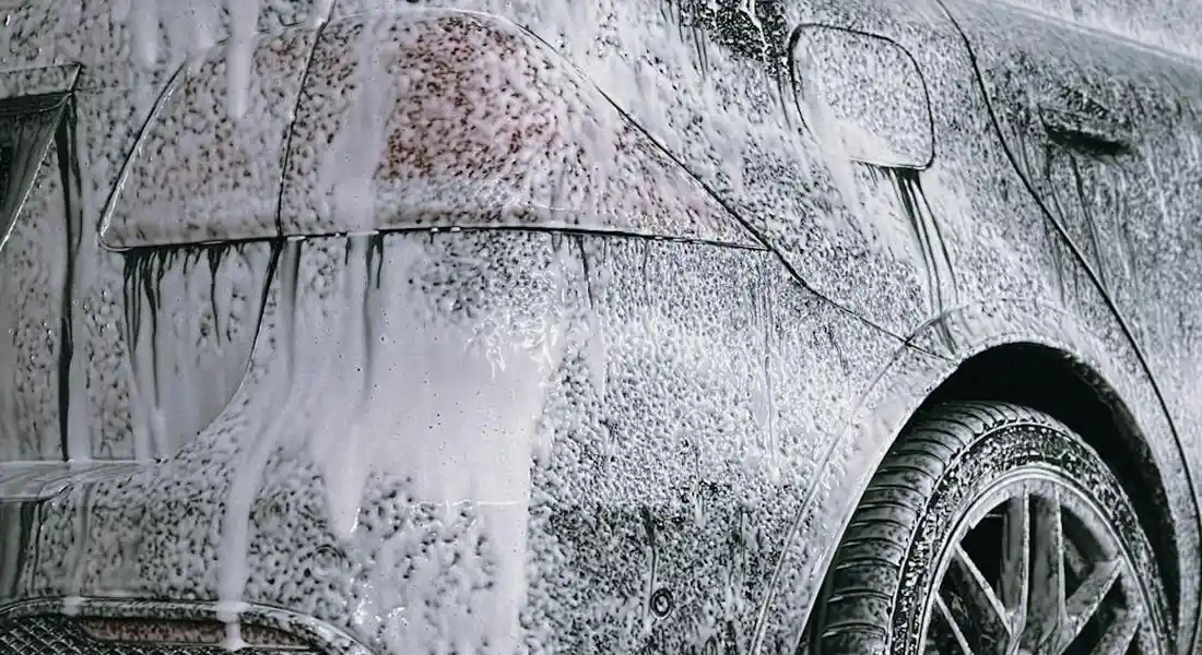 Side view of a car covered in foam and water during a wash, with the wheel visible.