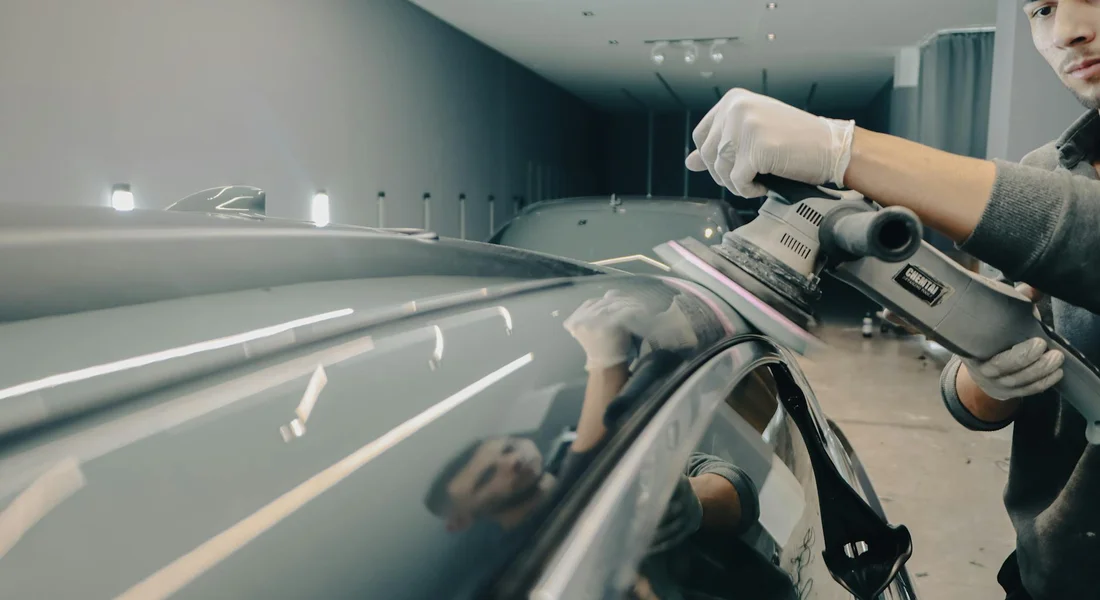 Detailer wearing gloves polishes a car panel with a power buffer in a well-lit workshop.