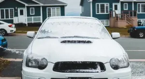 Front view of a white car covered in soap suds being washed on a residential street with houses in the background.