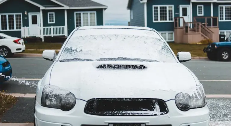 Front view of a white car covered in soap suds being washed on a residential street with houses in the background.