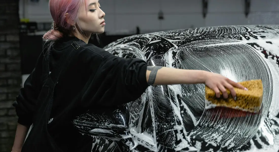 Person with pink hair using a sponge to wash a car's exterior windshield, creating foam on the glass.