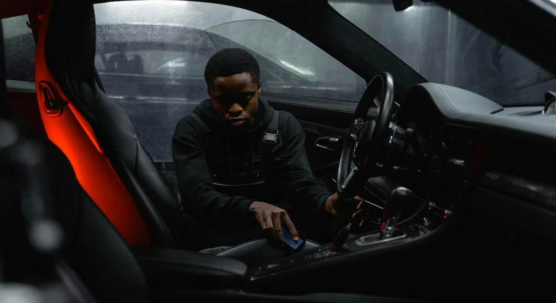 Inside a dark car, a person sits in the driver's seat and cleans the interior surfaces with a blue cloth near the center console.