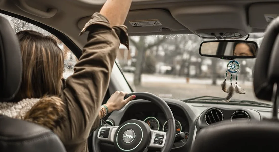 Inside a car cabin showing a driver in the front seat, with the dashboard and steering wheel in view; a dreamcatcher hangs from the rearview mirror.
