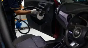 A person uses a spray bottle to clean fabric car seats and interior upholstery inside an open car door