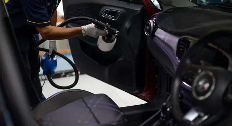 A technician wearing gloves sprays cleaning solution inside a car's interior door panel to treat mold and mildew on seats and carpeting.