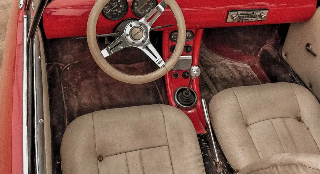 Interior of a vintage red car with tan cloth seats and a beige steering wheel, showing dirty floor mats.