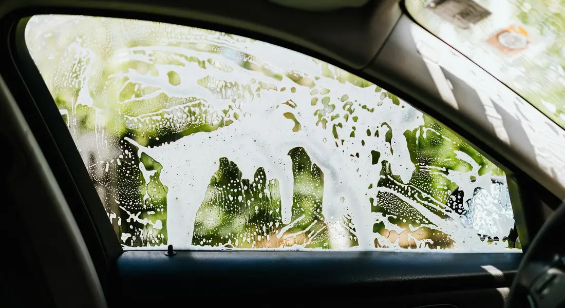 Car interior window covered in soap foam during cleaning, illustrating the preparation step before applying odor neutralizers to remove mildew and stale odors.