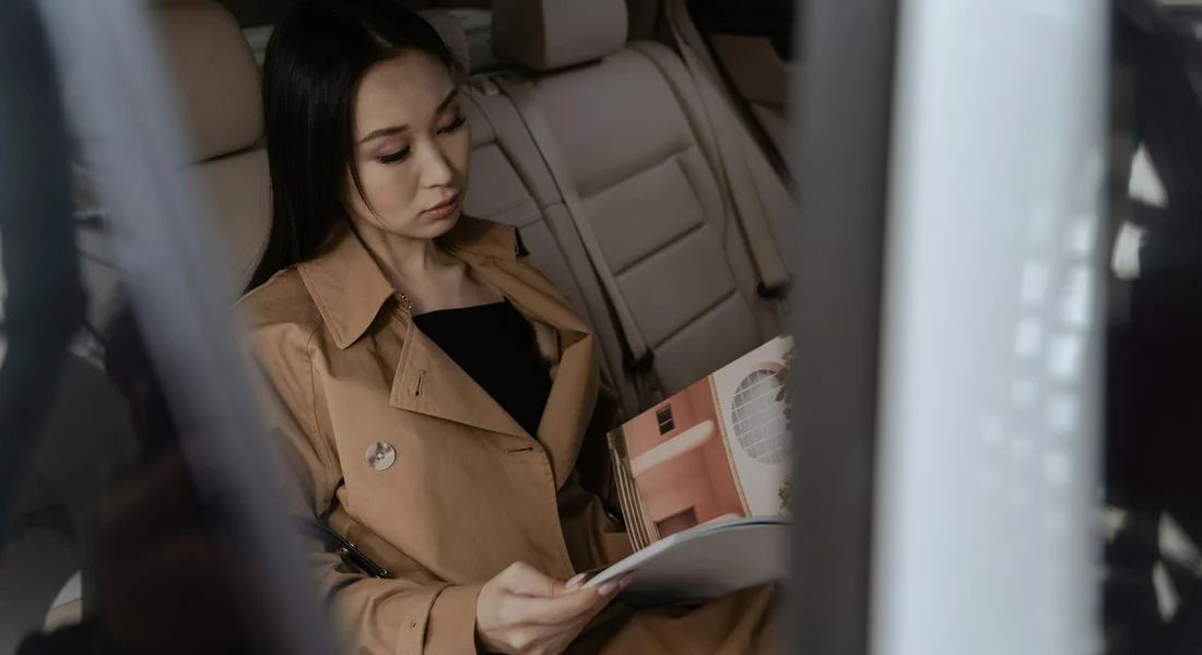 A woman in a tan coat sits in the back seat of a car, reading a magazine.