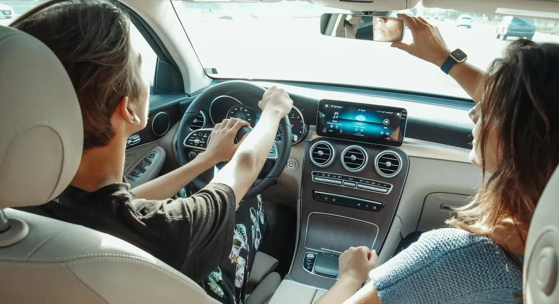 Interior view of a car dashboard and center console with two passengers; plastic dash and trim surfaces are visible.