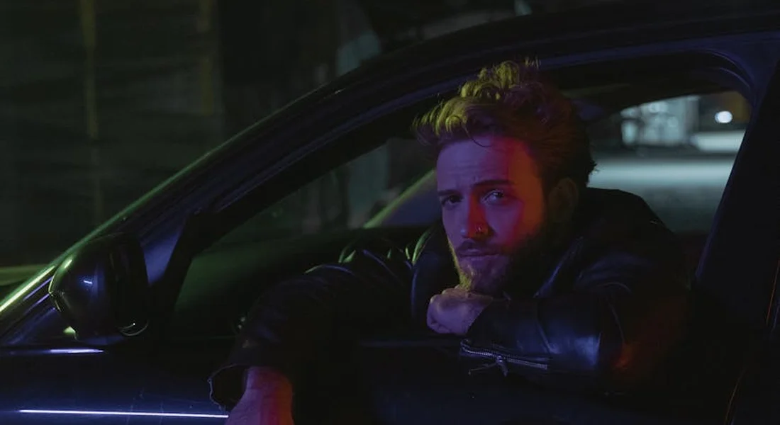 A man sits in a car at night, leaning with his chin on his hand, illuminated by blue and purple lighting.