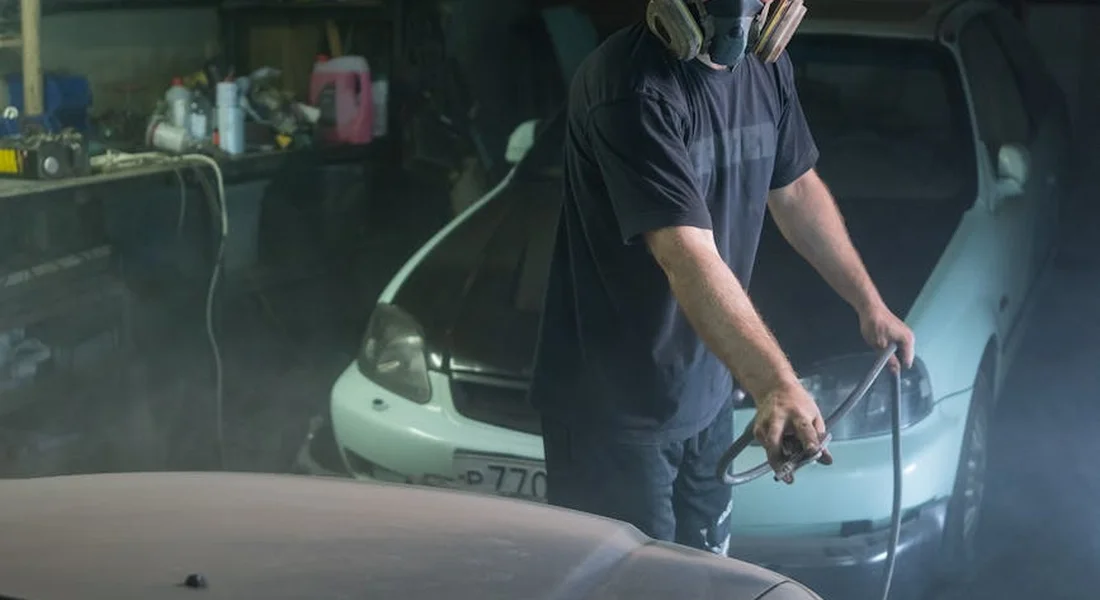Technician in a workshop wearing a mask and gloves works on a car, prepping the surface to prevent paint peeling on body panels.