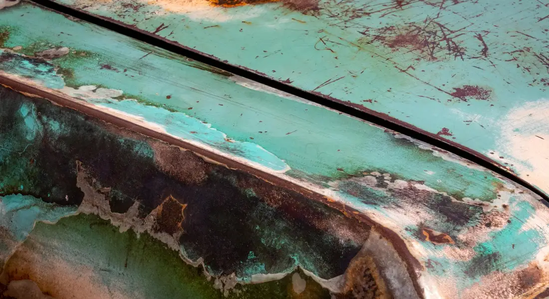 Close-up of a turquoise car panel with peeling paint and exposed corrosion, illustrating paint transfer and scraping marks.