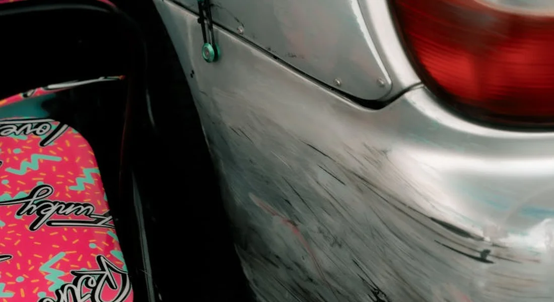 Close-up of a silver car door with light scratches and swirl marks, seen next to a pink graphic skateboard.