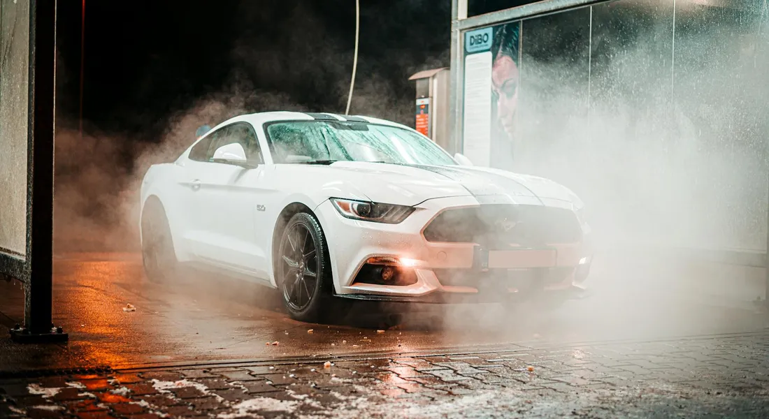 A white sports car being washed at a car wash with steam and spray, removing road salt from winter roads.
