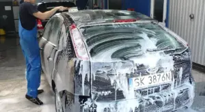 Technician applying foam wax to a dark hatchback in a workshop, with soap suds covering the rear of the car.