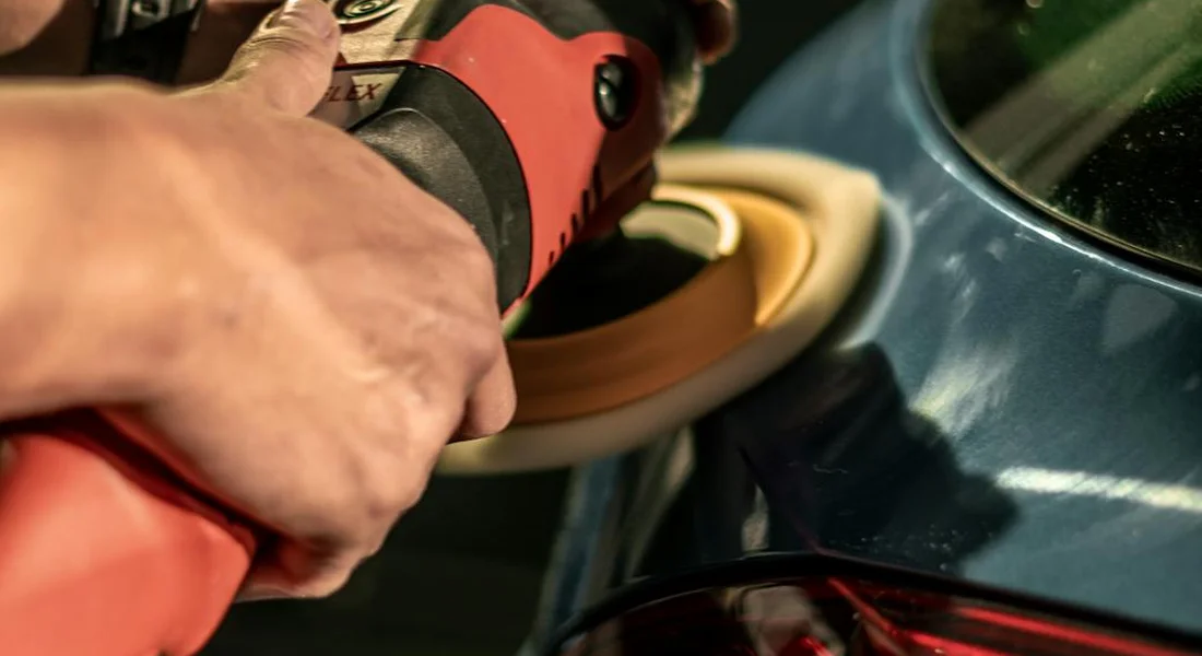 Close-up of a hand using an electric polisher on a blue car's surface to apply wax.