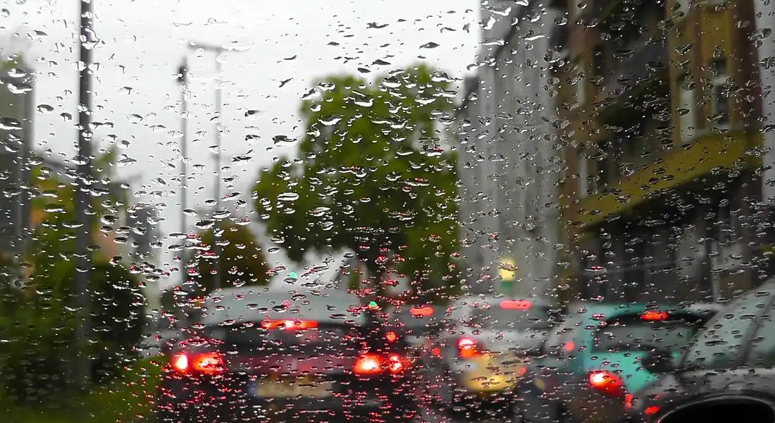 Rain droplets on a car windshield with blurred red brake lights and buildings in the background.