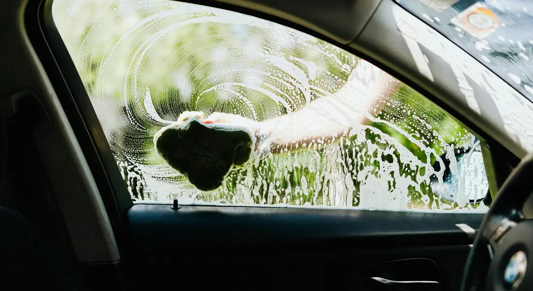 Outside view of a car window being cleaned; paint overspray on the glass with a gloved hand scrubbing and soapy streaks.