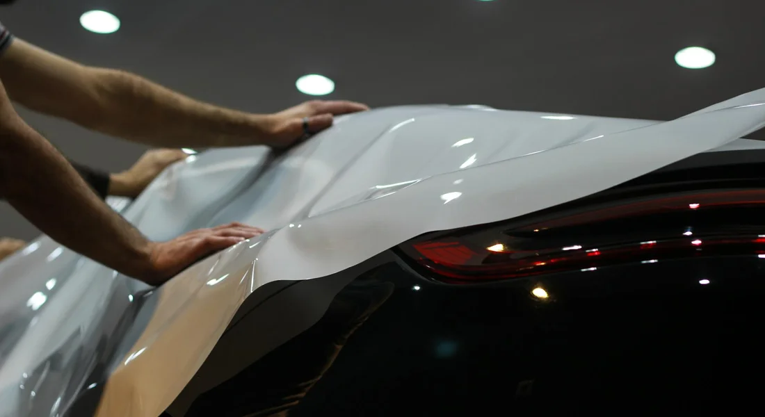 Hands smoothing a glossy vinyl wrap onto the curved surface of a car in a workshop.