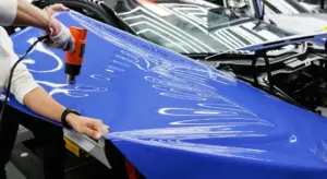 Technician applying blue vinyl wrap to a car hood using a heat gun.