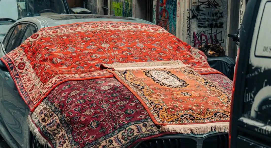 A car parked on a city street is covered with a colorful patterned rug, illustrating non-standard car covers.