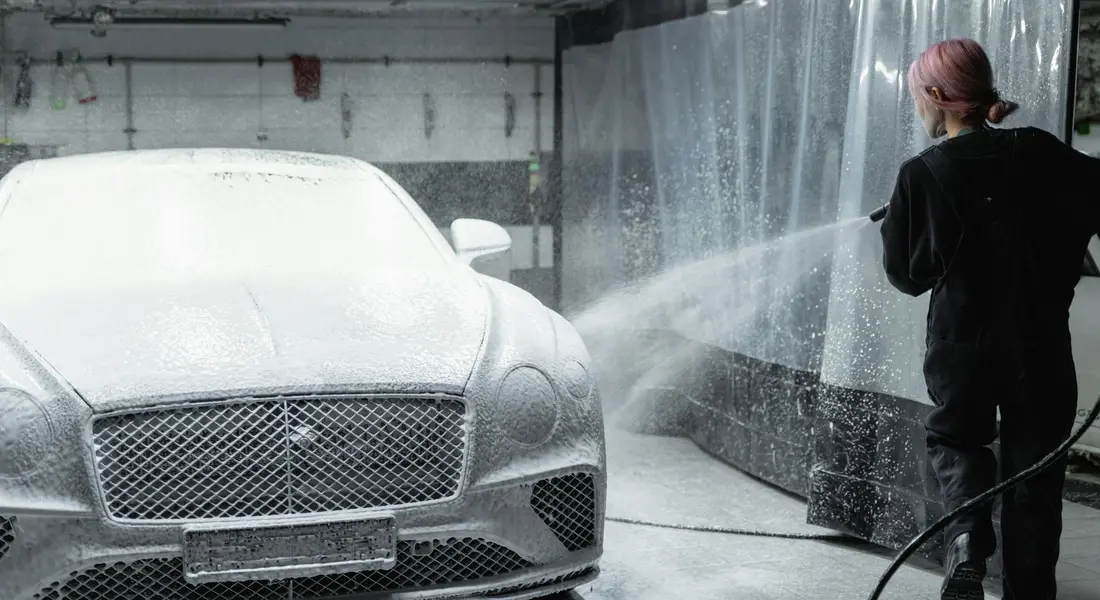 A car detailer uses a high-pressure washer to clean a luxury sedan inside a detailing bay.