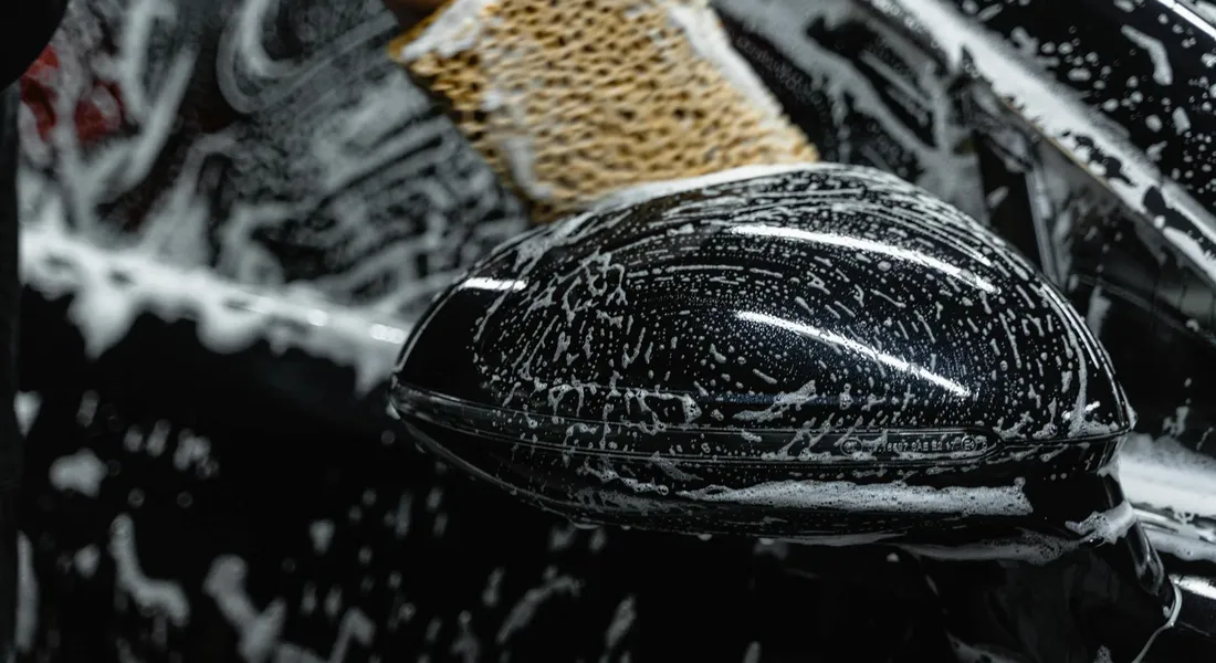 Close-up of a car's exterior side mirror and adjacent surface covered in soap suds during washing.