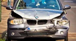 Front view of a silver BMW with a severely crumpled hood, damaged bumper, and broken headlight