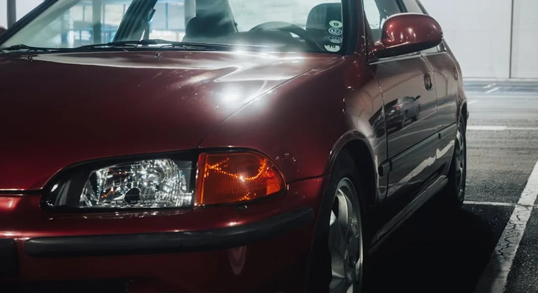 Close-up view of a maroon car in a garage, shown from the front-left side with headlight and bumper visible.