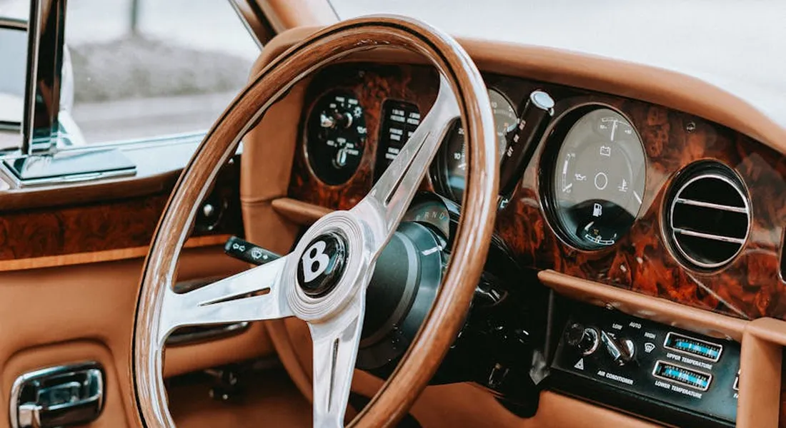 Interior of a luxury car with tan leather and wood trim, focusing on a chrome steering wheel with a Bentley emblem.