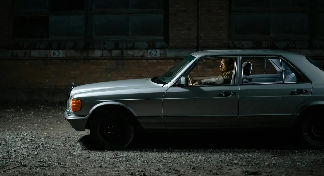 A person sits in the driver's seat of a silver sedan in a dimly lit outdoor area, capturing the moment a fuel odor may be noticed inside the car.