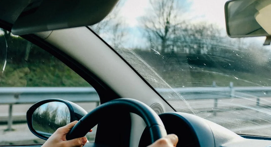Hands on the steering wheel inside a car, with the windshield and dashboard visible and a road scene outside.