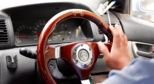 Interior of a car with a driver’s hand on the steering wheel, dashboard and windshield visible.