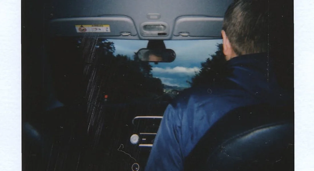 Interior of a car viewed from the back seat; a person wearing a blue jacket sits on the right, facing the front windshield, which shows smudges and grime with trees visible outside.