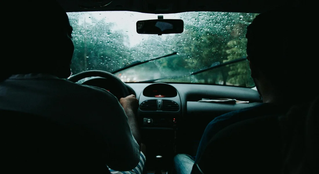 Interior of a car with two occupants sitting in the front seats; rain on the windshield with wipers clearing the glass.