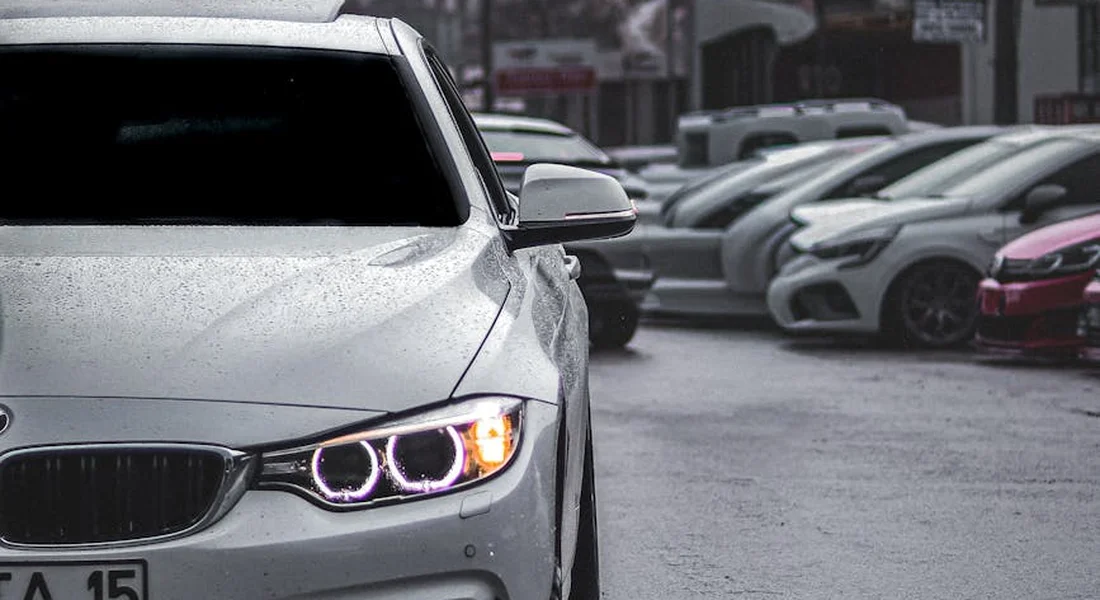 White sedan in a rainy parking lot, close-up of the front end with glowing headlights and other vehicles blurred in the background.