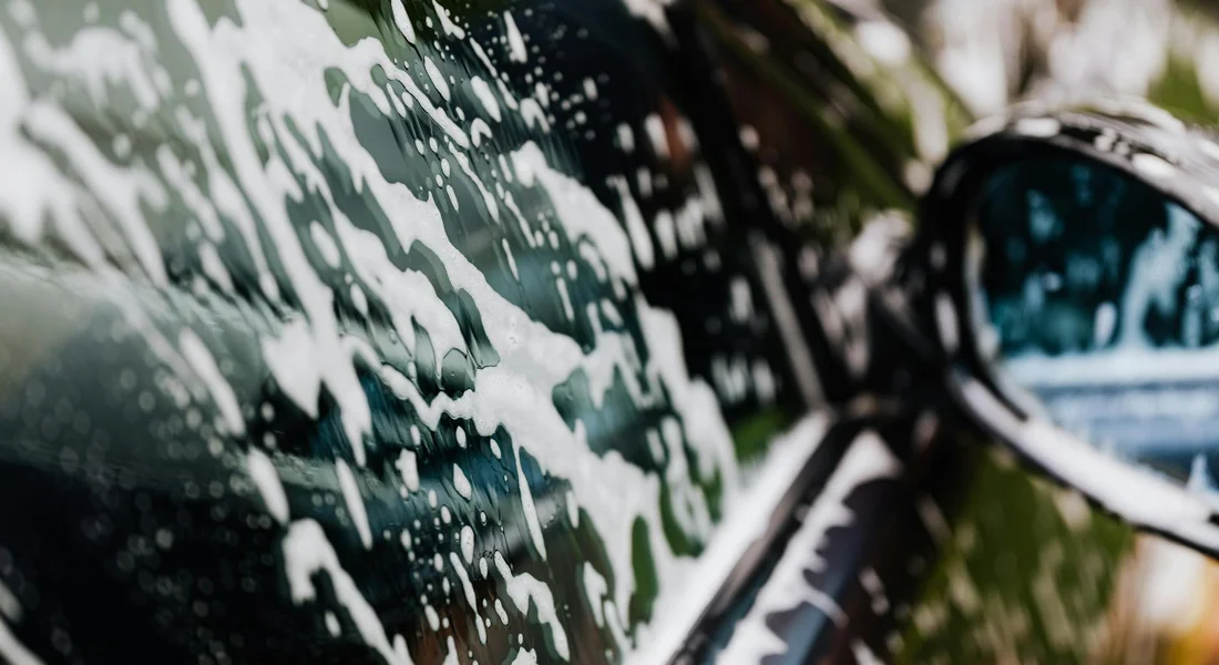 Close-up of a car's painted surface with soap suds during washing, with a side mirror visible in the background.