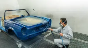 A technician wearing a protective suit and mask sprays blue automotive paint onto a car body inside an enclosed spray booth.